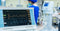 An EKG monitor displays heart activity in a medical office, with a doctor standing in the background.