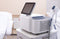 A laser hair removal machine with a digital screen stands beside a medical table in a clean and modern medical office.
