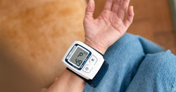 A person sits with a small blood pressure monitor on their wrist. The screen displays the letter "P" and the number "49."