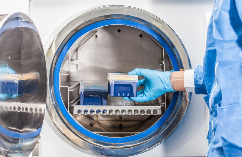 A person wearing a lab gown and gloves places items into a front-loading autoclave sterilizer.