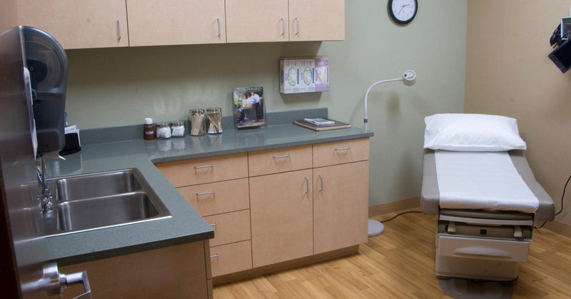 An electric exam table positioned in the corner of a medical examination room. A pillow and white paper are on the exam table.