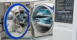 An autoclave sits on a white table. The door is open to reveal medical instruments in blue sterilization pouches.