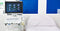A portable ultrasound machine displays images on the screen next to a hospital bed in front of a blue wall.