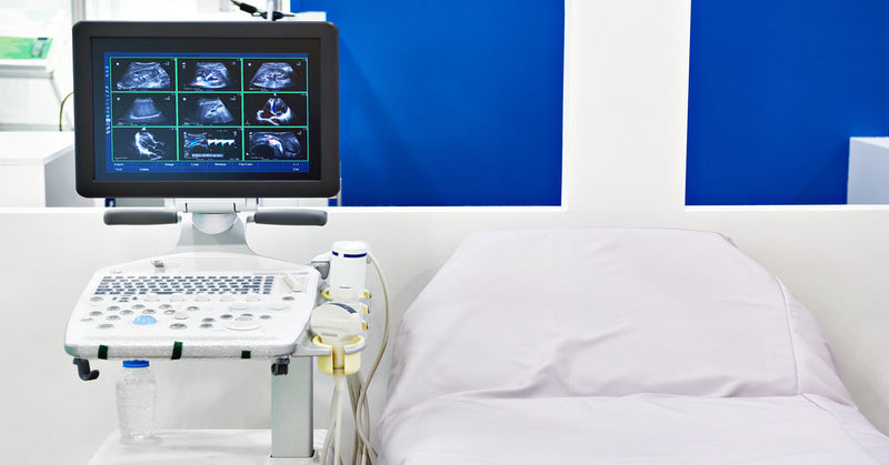 A portable ultrasound machine displays images on the screen next to a hospital bed in front of a blue wall.