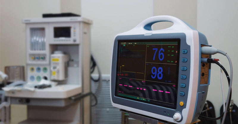 A close-up shows an ECG/EKG heart rate monitor sitting in a medical office, displaying a reading of 76 over 98.