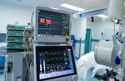 Two medical monitors display a patient's vital signs in a hospital room, with more medical equipment in the background.