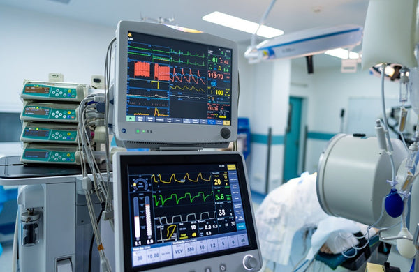 Two medical monitors display a patient's vital signs in a hospital room, with more medical equipment in the background.