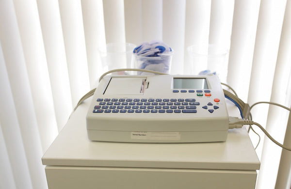 An ECG machine with blue buttons and various wires sits on a cabinet, near a window with white curtains.