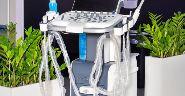 A portable ultrasound machine with display screens, a control panel, and multiple probes on a wheeled base in a medical setting.