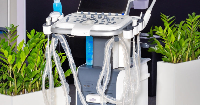 A portable ultrasound machine with display screens, a control panel, and multiple probes on a wheeled base in a medical setting.