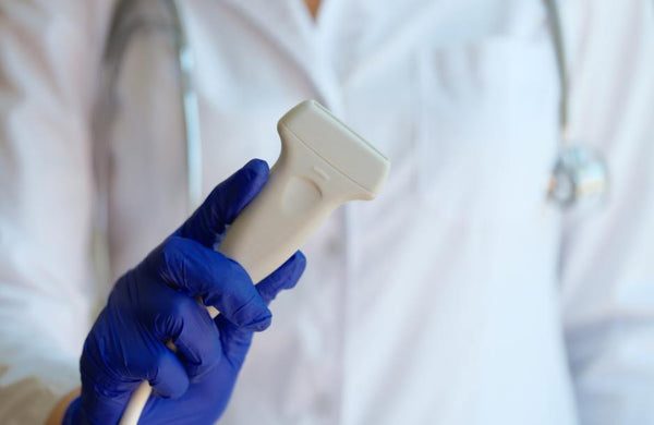A medical professional in a white lab coat and blue gloves holds an ultrasound Doppler in a clinical setting.